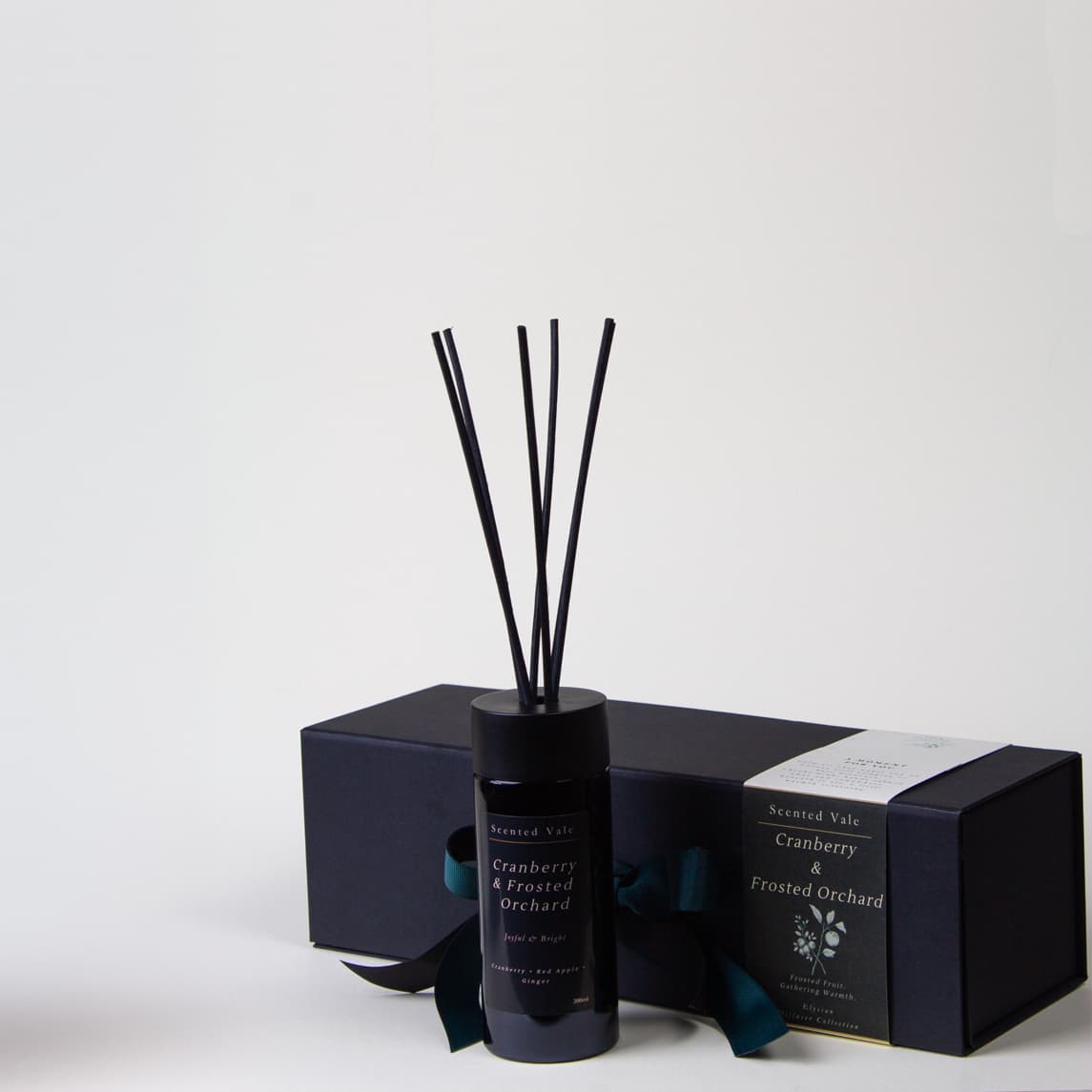 A reed diffuser bottle with black reeds and a violet glass bottle, shown with its matching black gift box and paper band on a clean white background. The label reads 'Scented Vale Cranberry & Frosted Orchard'.