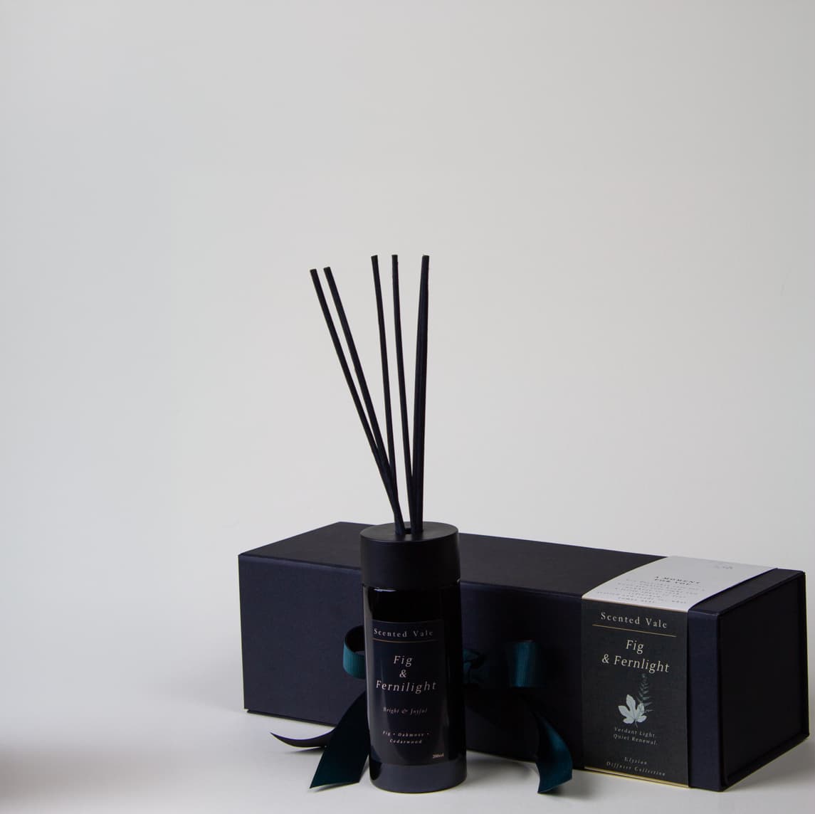 A reed diffuser bottle with black reeds stands in front of a dark presentation box with a teal ribbon, on a clean light grey background.