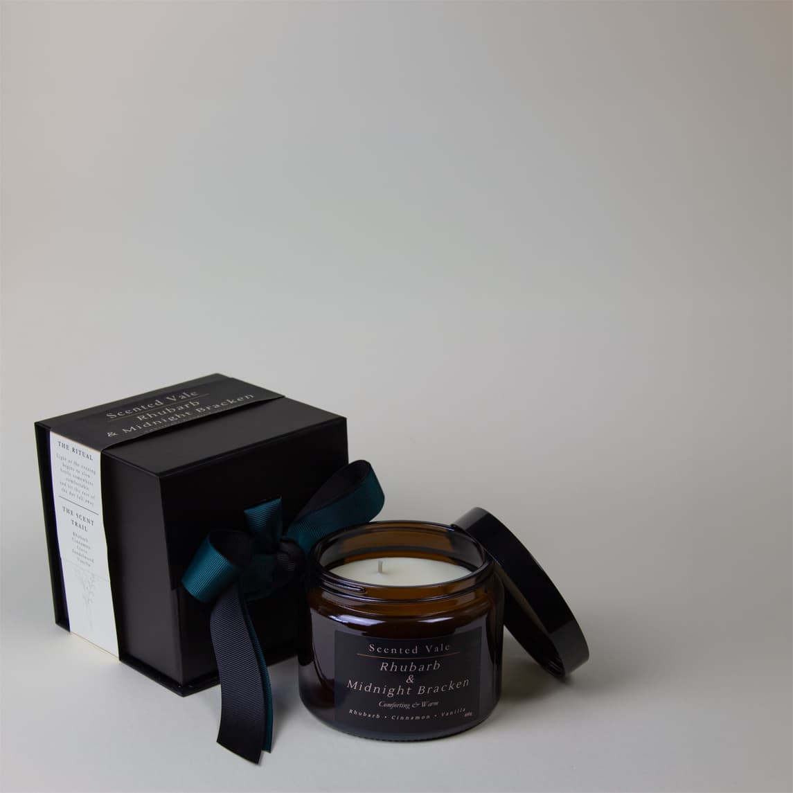 An amber glass jar candle with its black lid beside a black presentation box wrapped in a dark teal ribbon. The candle label reads Scented Vale Rhubarb & Midnight Bracken.