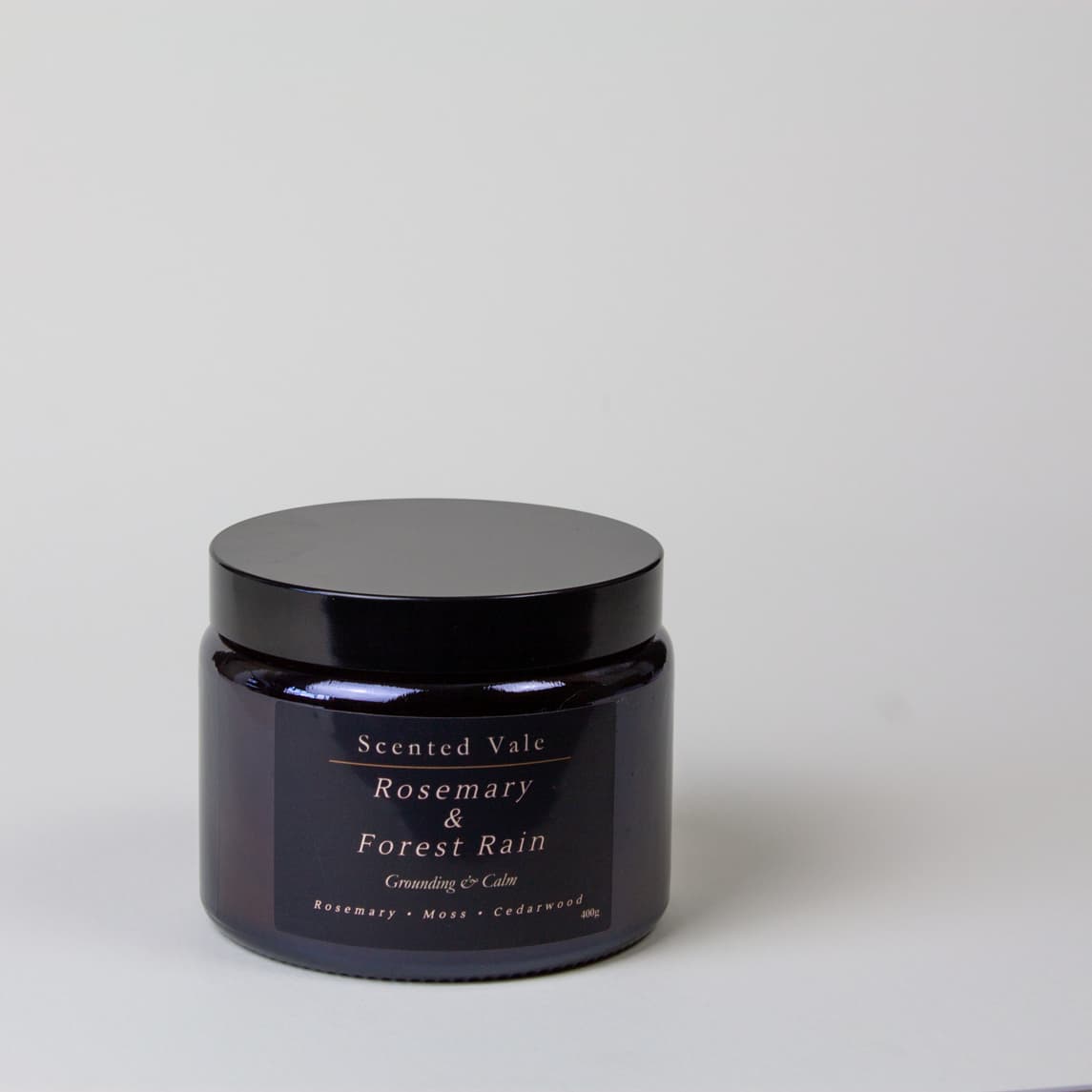 A dark amber glass jar with a black lid, labeled 'Scented Vale Rosemary & Forest Rain', on a light grey background.