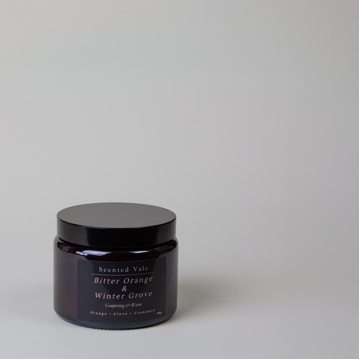 A dark amber glass jar candle with a black lid and a black label reading 'Scented Vale Bitter Orange & Winter Grove' on a light grey background.
