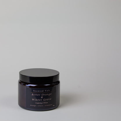 A dark amber glass jar candle with a black lid and a black label reading 'Scented Vale Bitter Orange & Winter Grove' on a light grey background.