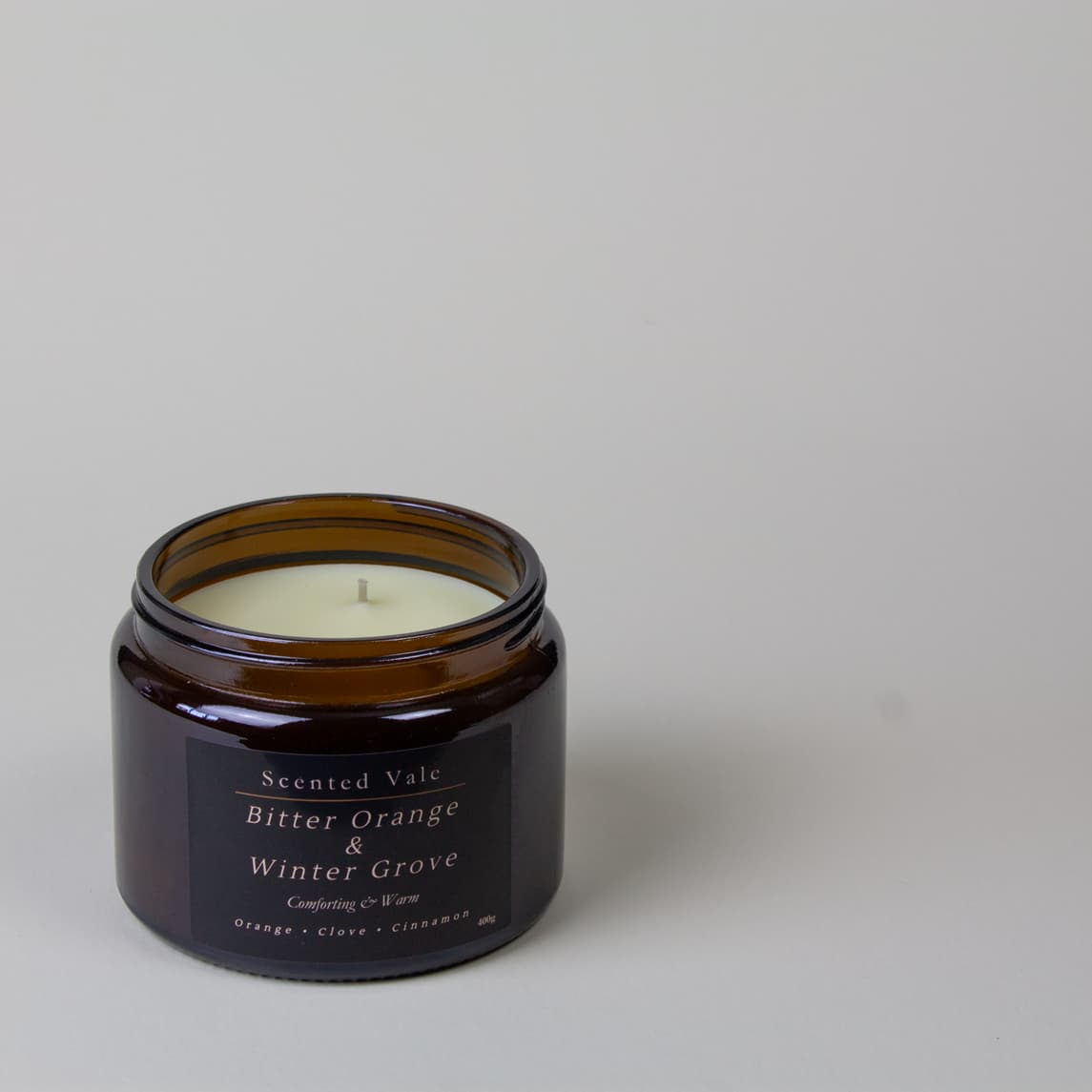 An amber glass jar candle with a black label reading 'Scented Vale Bitter Orange & Winter Grove' on a light grey background.