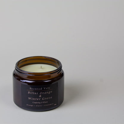 An amber glass jar candle with a black label reading 'Scented Vale Bitter Orange & Winter Grove' on a light grey background.