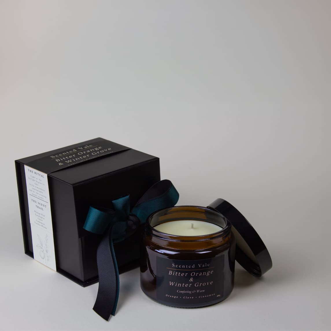 An amber glass jar candle with its lid off, shown next to a black gift box tied with a dark teal ribbon. The candle label reads 'Scented Vale Bitter Orange & Winter Grove'.