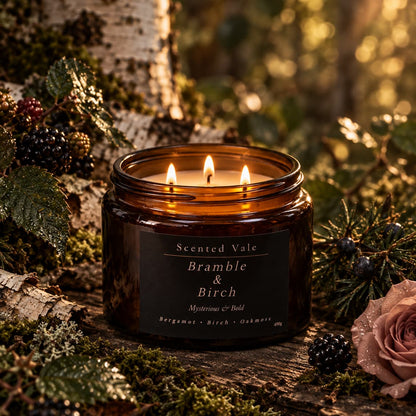 A lit amber glass jar candle labeled 'Scented Vale Bramble & Birch' sits on a mossy log outdoors, surrounded by berries, evergreen branches, and soft forest light.