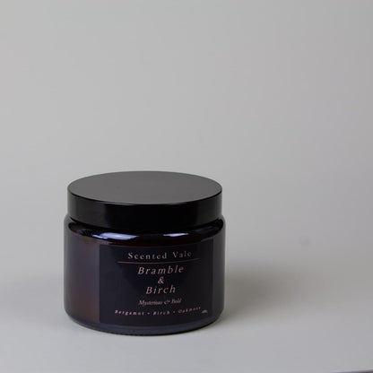 A dark amber glass jar candle with a black lid and a black label reading 'Scented Vale Bramble & Birch' on a light grey background.