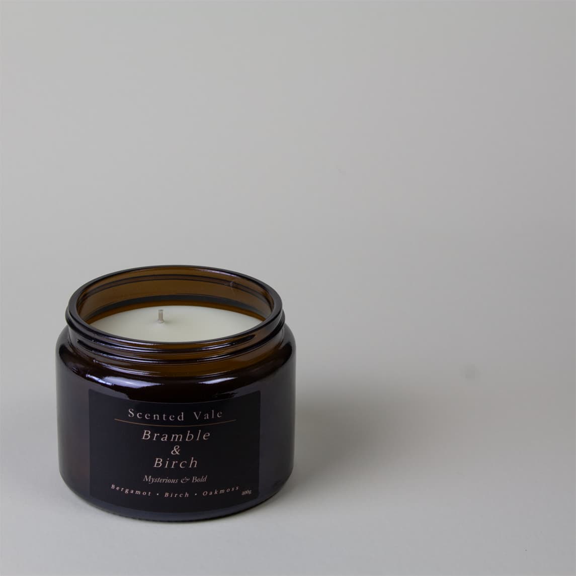 An amber glass jar candle with white wax and a black label reading 'Scented Vale Bramble & Birch' on a light grey background.