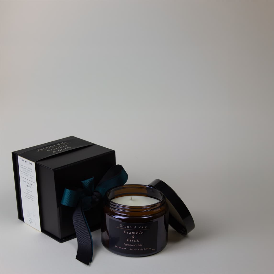 A scented candle in an amber glass jar with its lid off, placed beside a black gift box tied with a dark ribbon. The candle label reads 'Scented Vials' and 'Bramble Birch'.