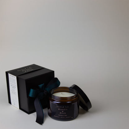 A scented candle in an amber glass jar with its lid off, placed beside a black gift box tied with a dark ribbon. The candle label reads 'Scented Vials' and 'Bramble Birch'.