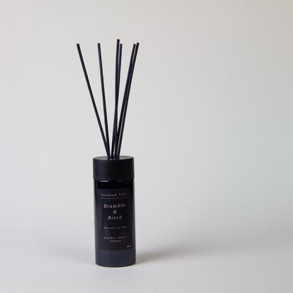 A reed diffuser bottle with black reeds stands on a plain grey background. The label reads 'Scented Vale Bramble & Birch'
