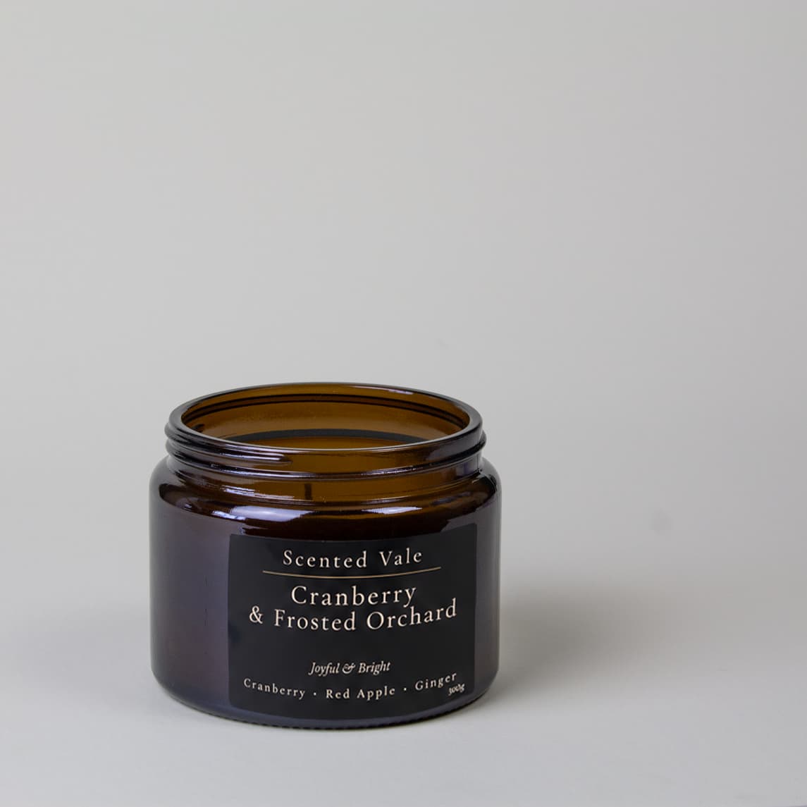 An amber glass jar candle with a black label reading 'Scented Vale Cranberry & Frosted Orchard' on a light gray background.