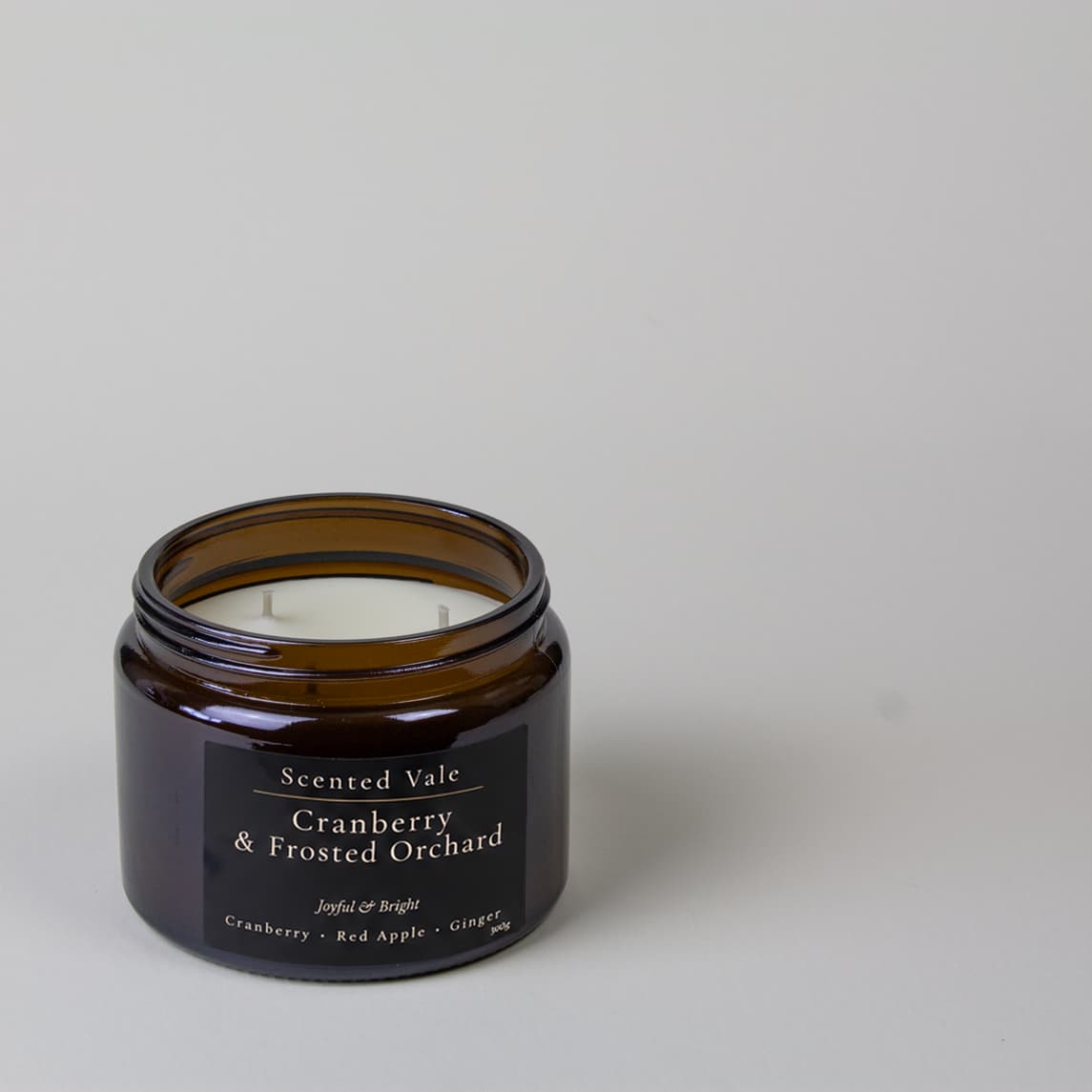An amber glass jar candle with a black label reading 'Scented Vale Cranberry & Frosted Orchard' on a light gray background.