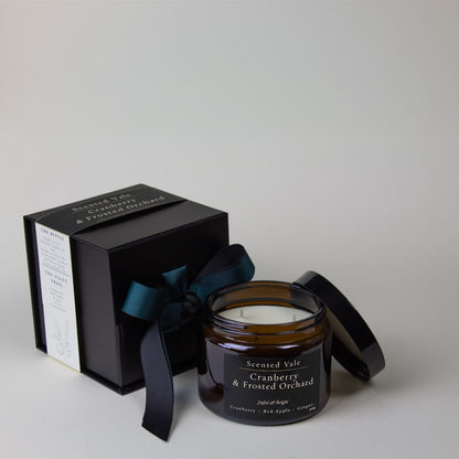 An amber glass jar candle with a black lid and label reading 'Scented Vale Cranberry & Frosted Orchard,' shown beside a black gift box tied with a dark ribbon on a light gray background.