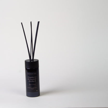 A reed diffuser bottle in a violet glass container with six dark reeds, labelled 'Cranberries & Frosted Orchard', set against a plain light grey background.