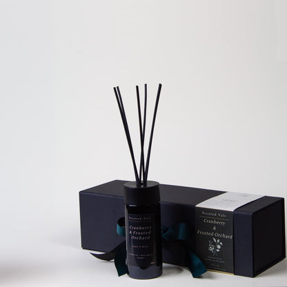 A reed diffuser bottle with black reeds and a violet glass bottle, shown with its matching black gift box and paper band on a clean white background. The label reads 'Scented Vale Cranberry & Frosted Orchard'.