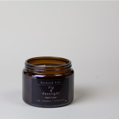 An amber glass jar candle with a black label reading 'Scented Vale Fig & Fernlight' on a light gray background.