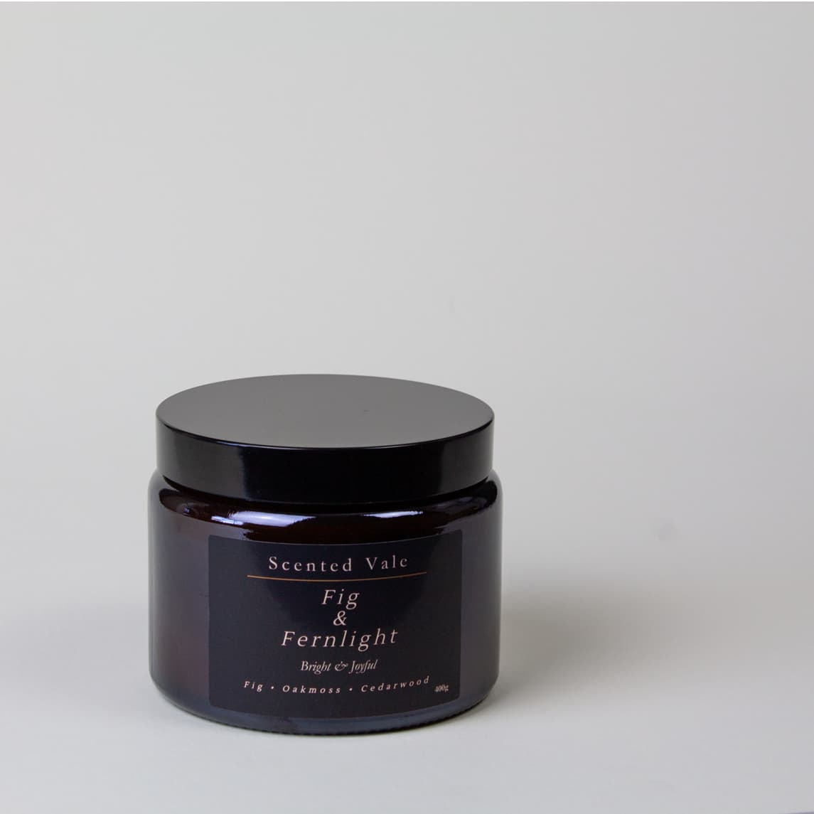 A dark amber glass jar with a black lid and a black label reading 'Scented Vale Fig & Fernlight' on a light gray background.