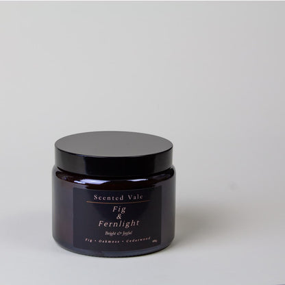 A dark amber glass jar with a black lid and a black label reading 'Scented Vale Fig & Fernlight' on a light gray background.
