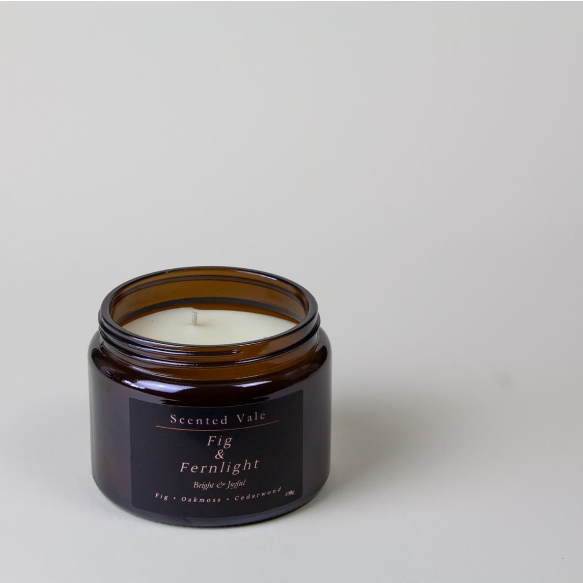 An amber glass jar candle with a white wax fill and a black label reading 'Scented Vale Fig & Fernlight', photographed on a clean light gray background.