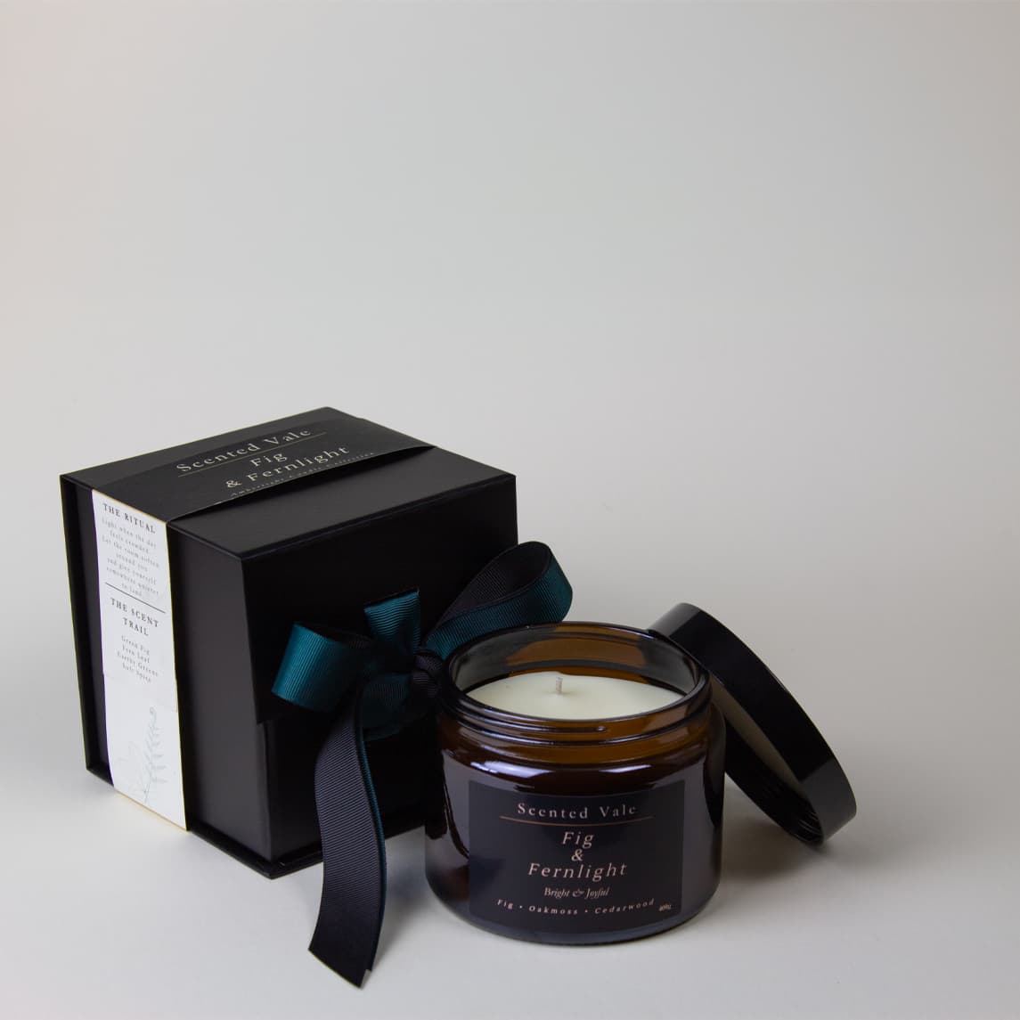 An amber glass jar candle with its black lid beside a black presentation box tied with a dark ribbon, on a clean light gray background. The labels read 'Scented Vale Fig & Fernlight'.