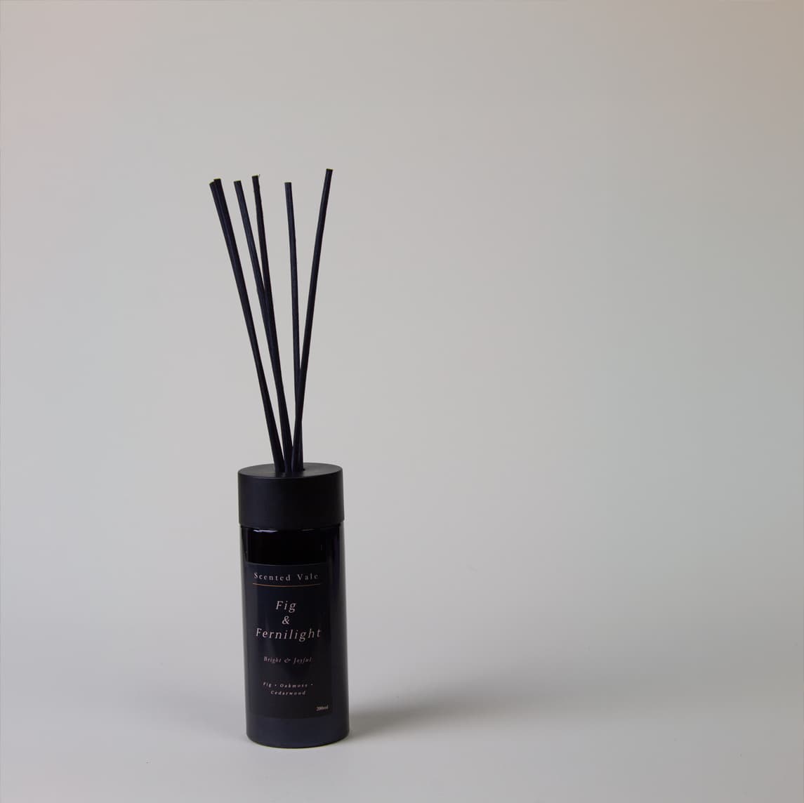 A reed diffuser bottle in matte black with six dark reeds, photographed against a clean light grey background. The label reads 'Scented vale' and 'Fig & Fernlight'.