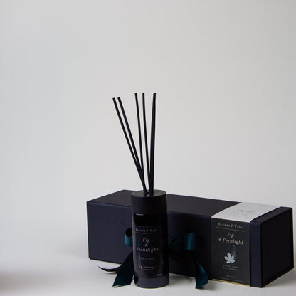 A reed diffuser bottle with black reeds stands in front of a dark presentation box with a teal ribbon, on a clean light grey background.