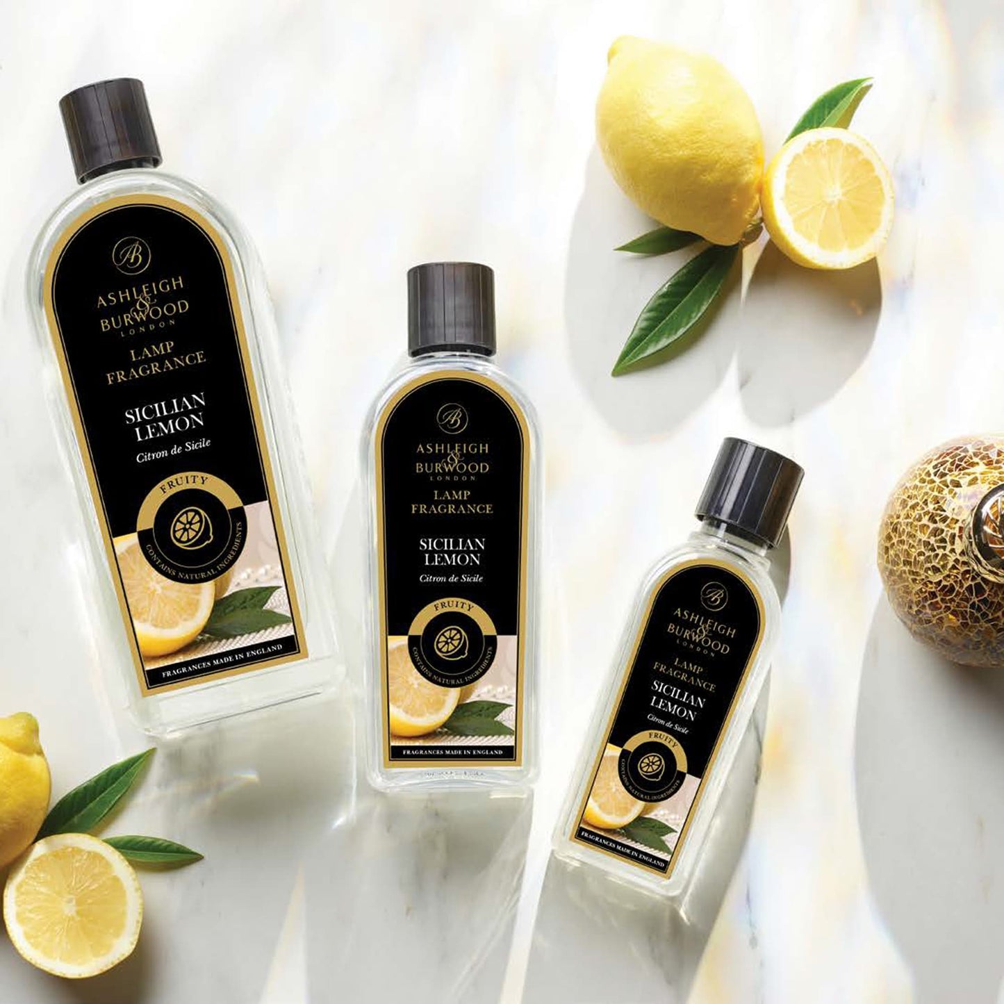 Three different sized bottle of Ashleigh & Burwood Sicilian Lemon Lamp Fragrance on a white marble board with whole and Sliced lemons