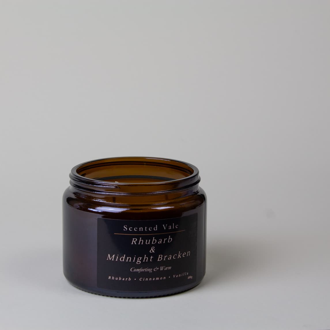 An amber glass jar candle with a black label reading, Scented Vale Rhubarb & Midnight Bracken, on a light grey background.