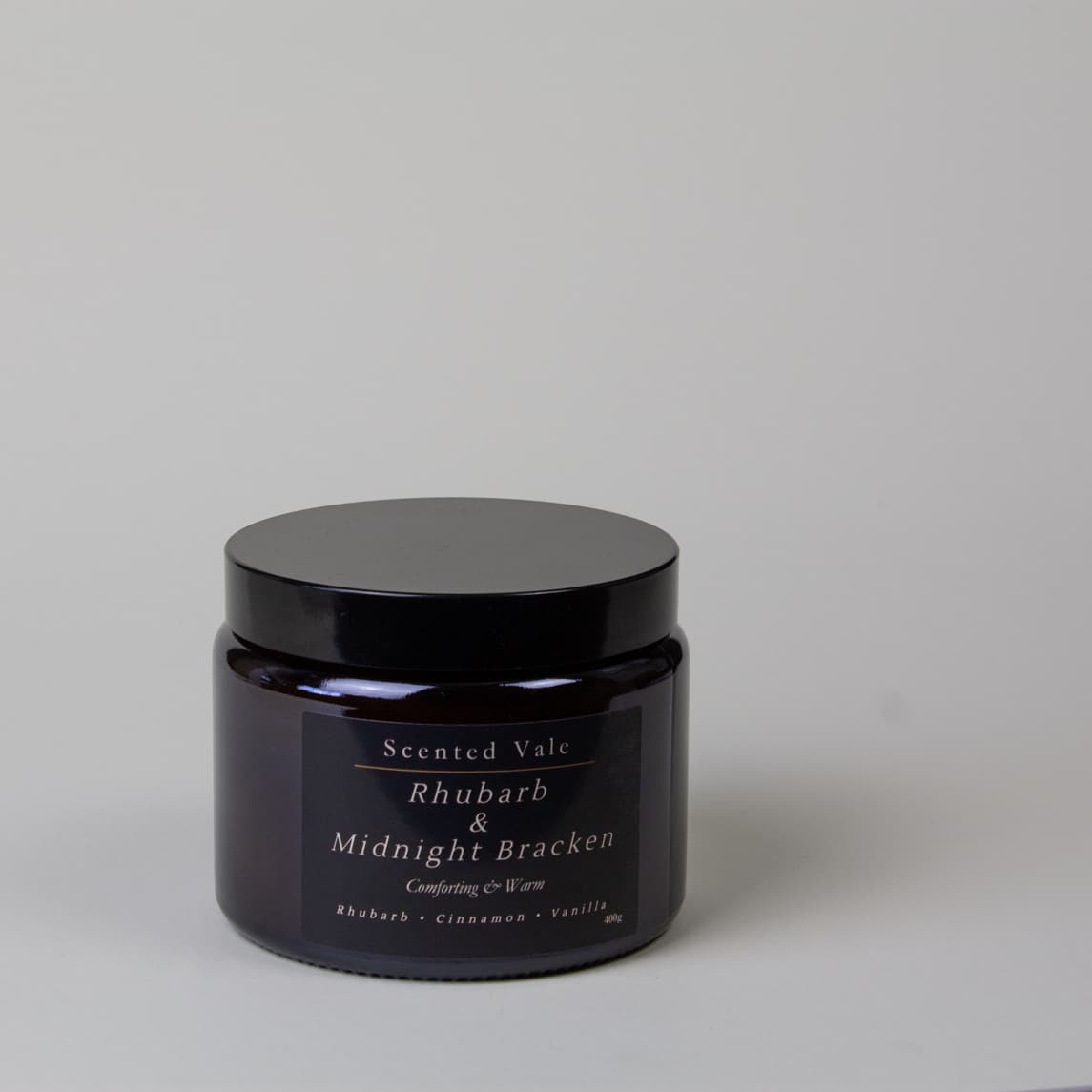 A dark amber glass jar with a black lid and a dark label reading, Scented Vale Rhubarb & Midnight Bracken, on a light grey background.