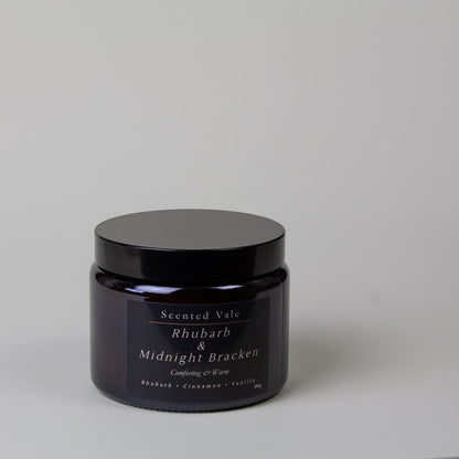 A dark amber glass jar with a black lid and a dark label reading, Scented Vale Rhubarb & Midnight Bracken, on a light grey background.