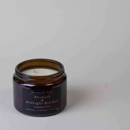 An amber glass jar candle with a white wax fill and a black label reading, Scented Vale Rhubarb & Midnight Bracken. The candle is photographed on a clean, light grey surface with a matching grey background.