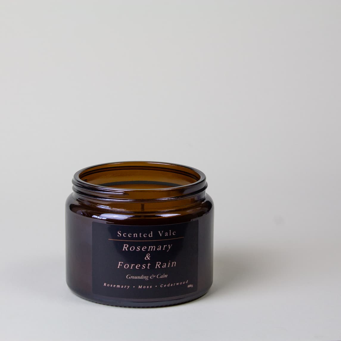 An amber glass jar candle with a black label reading 'Scented Vale Rosemary & Forest Rain' on a light grey background.