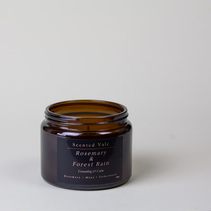 An amber glass jar candle with a black label reading 'Scented Vale Rosemary & Forest Rain' on a light grey background.