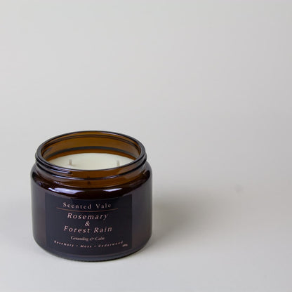 An amber glass jar candle with a black label reading 'Scented Vale Rosemary & Forest Rain' on a light grey background.