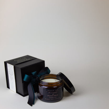 An amber glass jar candle with a black lid beside a black gift box tied with a dark teal ribbon. The candle label reads 'Scented Vale Rosemary & Forest Rain'