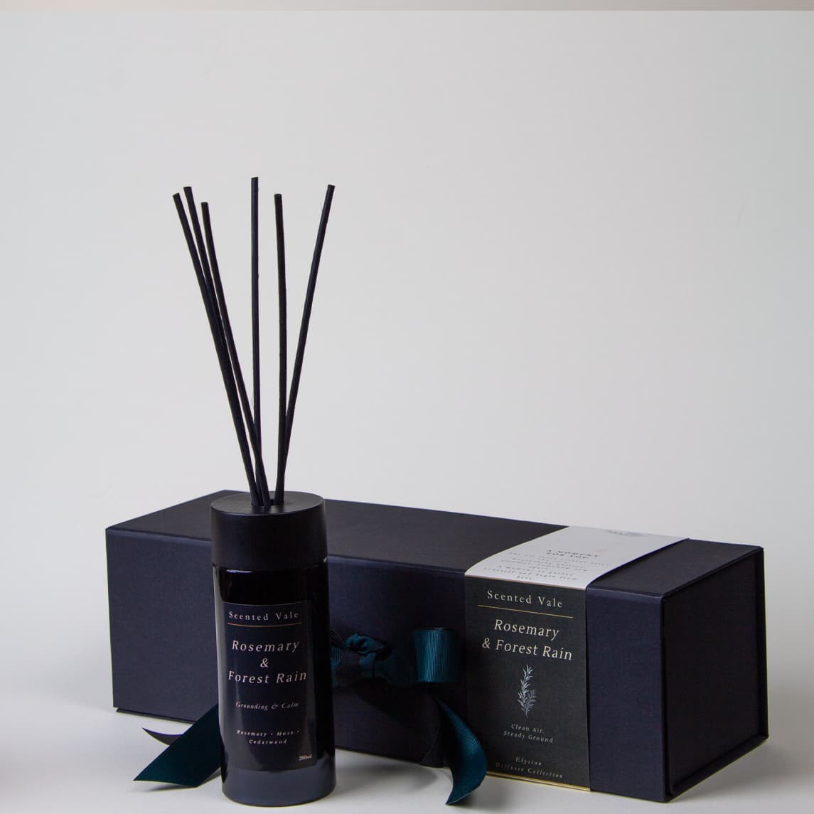 A reed diffuser bottle with black reeds stands in front of its matching dark gift box. The bottle and box are labeled 'Scented Vale Rosemary & Forest rain' with a dark teal ribbon tied around the box.