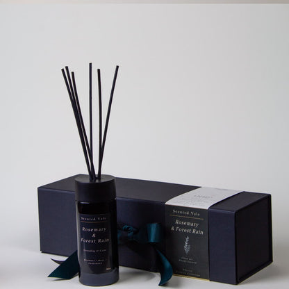 A reed diffuser bottle with black reeds stands in front of its matching dark gift box. The bottle and box are labeled 'Scented Vale Rosemary & Forest rain' with a dark teal ribbon tied around the box.