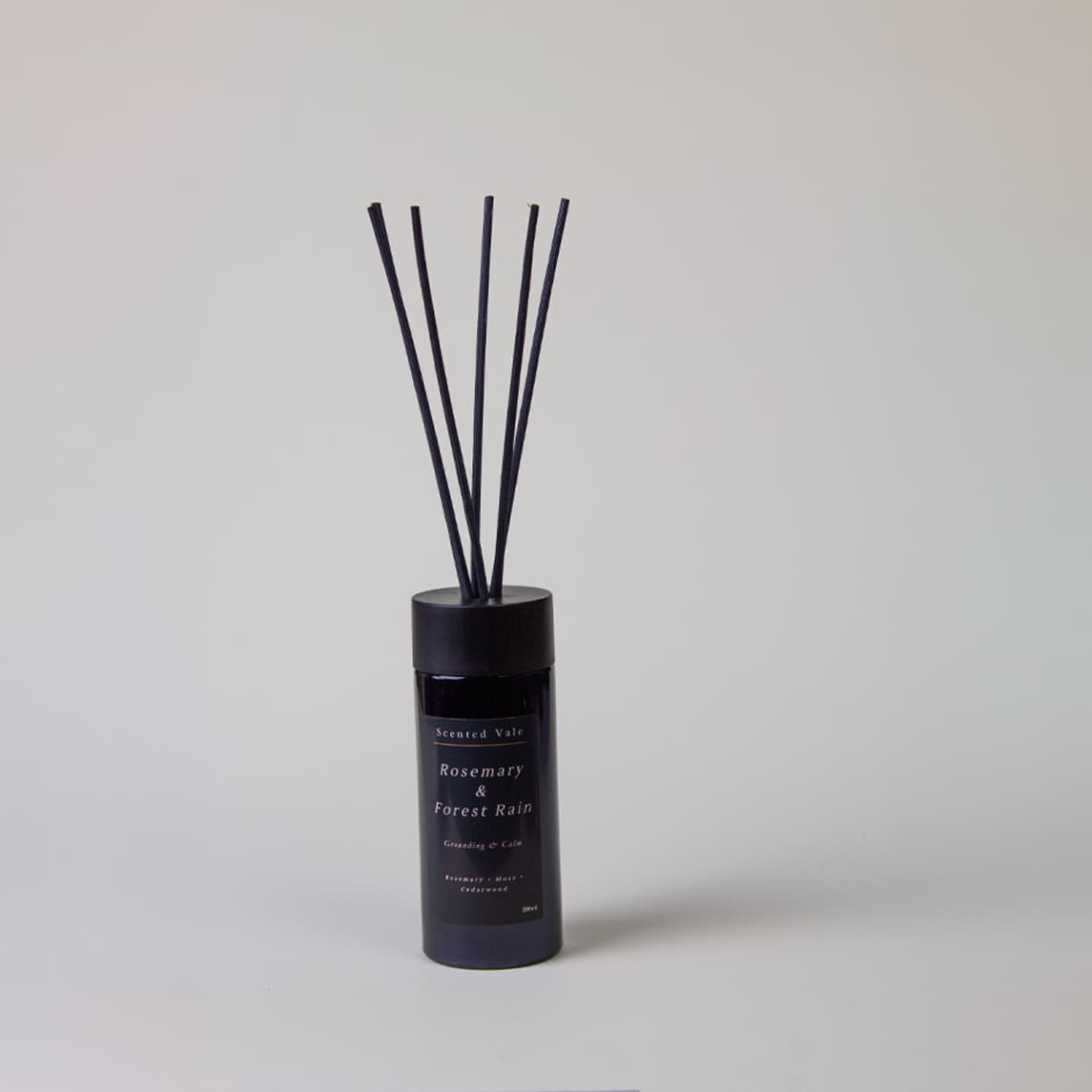 A reed diffuser bottle in a dark navy blue glass container with six black reeds, labeled 'Rosemary & Forest Rain' by 'Scented Vale', set against a plain light grey background.