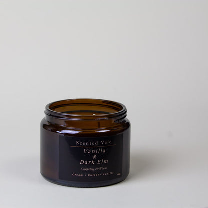 An amber glass jar candle with a black label reading 'Scented Vale Vanilla & Dark Elm' on a light grey background.