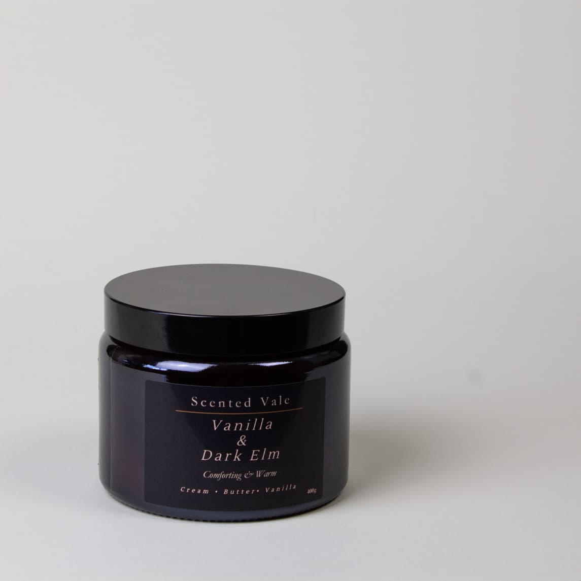 A dark amber glass jar candle with a black lid and a black label reading 'Scented Vale Vanilla & Dark Elm' on a light grey background.