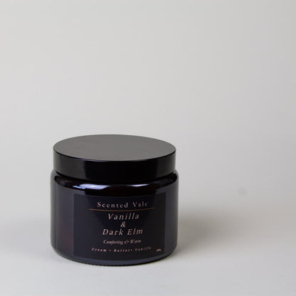 A dark amber glass jar candle with a black lid and a black label reading 'Scented Vale Vanilla & Dark Elm' on a light grey background.