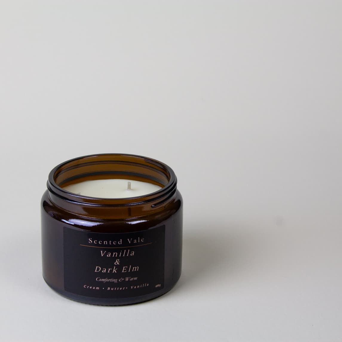 An open amber glass jar candle with white wax and a dark label reading 'Scented Vale Vanilla & Dark Elm' on a light grey background.