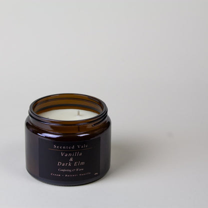 An open amber glass jar candle with white wax and a dark label reading 'Scented Vale Vanilla & Dark Elm' on a light grey background.
