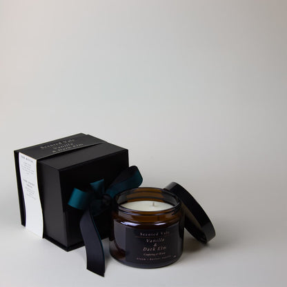 An amber glass jar candle with its lid off, next to a black presentation box tied with a dark teal ribbon. The labels read Scented Vale Vanilla & Dark Elm.