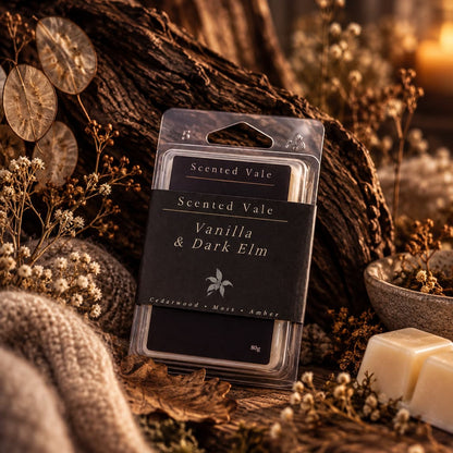 Scented Vale Vanilla & Dark Elm wax melts in a clear plastic clamshell package with a dark label, styled on a rustic wood surface with dried flowers, a knitted textile, and two cream wax bars nearby.